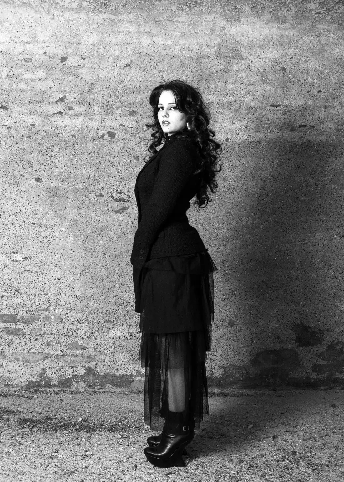 Costina Cindea, full length in a black tailored jacket and tulle skirt, looking back over her shoulder against a concrete wall.